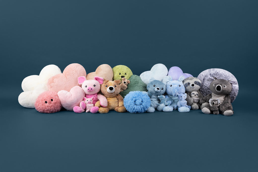 This image features a lineup of Hugimals World products styled as a rainbow, with different colored Hugimals Plushies, pillows, Hug Ball and Hug Babies in a horizontal array featuring from left to right the colors white, pink, tan, green, blue, lavender and gray. 