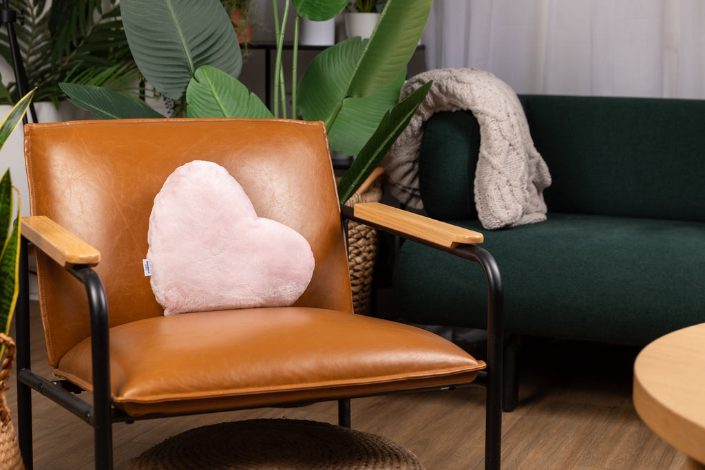 This image shows the pink Heart to Hug Pillow on a leather chair in a living room, with plants and a green couch behind the chair.