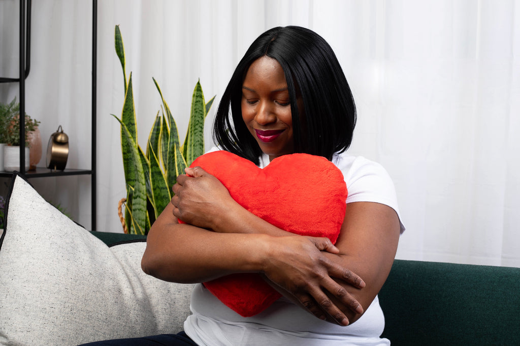 This image shows a woman holding a red heart-shaped pillow close to her chest, smiling with her eyes closed. She's sitting on a dark green couch and wearing a white t-shirt. 