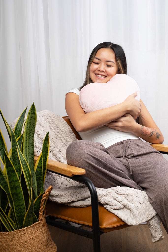 This image shows a smiling young woman with oat-colored pajama pants and a white t-shirt sitting comfortably on an arm chair and hugging a pink heart-shaped pillow under her face.
