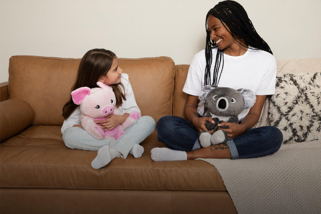 This image shows a little girl sitting on a couch holding Harper the Pig Hugimal. Next to her is a young woman holding Quinn the Koala Hugimal. They are both wearing white t-shirts and sitting cross-legged on the couch. They are smiling at each other