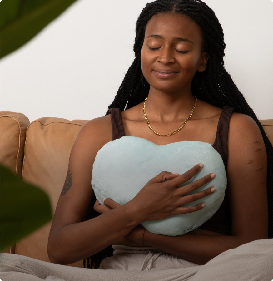 This photo shows a young woman (late teens/early 20s) holding a Mist Heart to Hug Pillow tight to her chest as she sits cross-legged on a tan couch. She is wearing light colored pajama pants and a black tank top with a gold necklace. Her eyes are closed and she is smiling slightly, looking comforted. She is on a tan couch and there is a blg plant showing in the foreground.