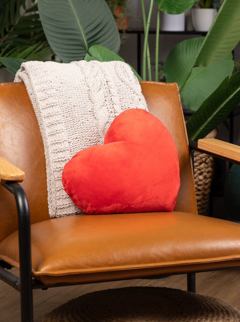 This image shows the red Heart to Hug Pillow placed on a tan leather chair that has a white blanket draped over it. There i a green plant behind the chair.