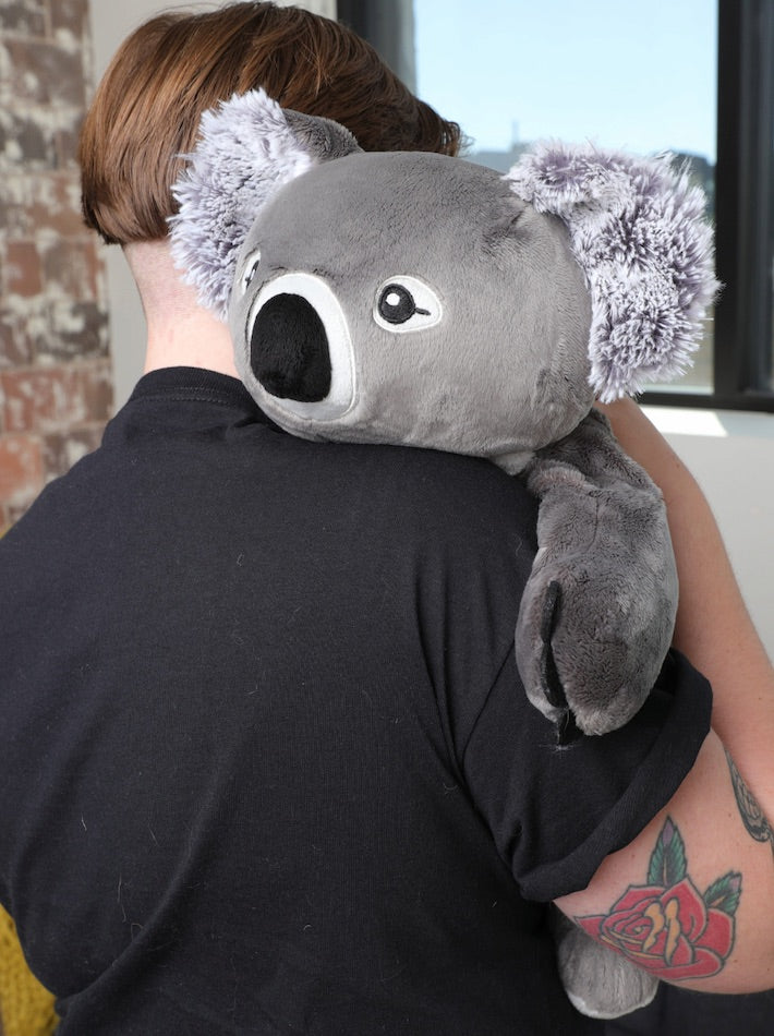 A woman with short hair, tattoos, and a black t-shirt is faced away from us, while a koala stuffed animal is hugging her.