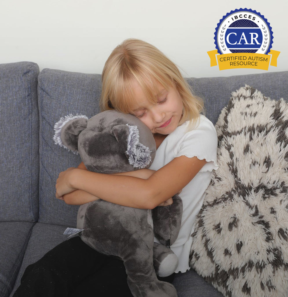 This image shows a little blond girl (6 or 7 years old) wearing a white tee shirt and black pants, sitting on a gray couch against a white and gray pillow. The girl is hugging Quinn the Koala Hugimal tight with her eyes closed, looking comforted. On the top right is a seal that says Certified Autism Resource, a distinction awarded by IBCCES.