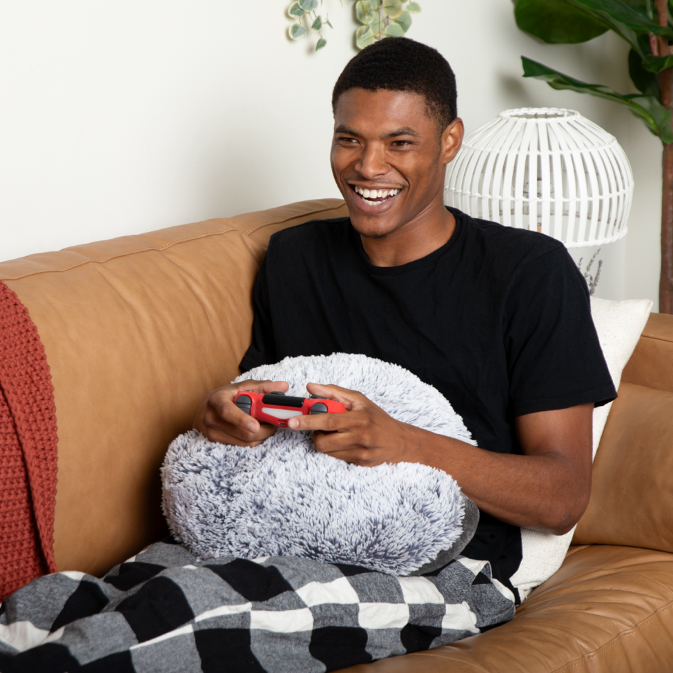 This photo shows a young man (early to mid 20s) on a tan couch smiling, holding a video game console, with a Hug Pillow (round and gray) on his lap. He is wearing plaid pajama pants and a black tee shirt.
