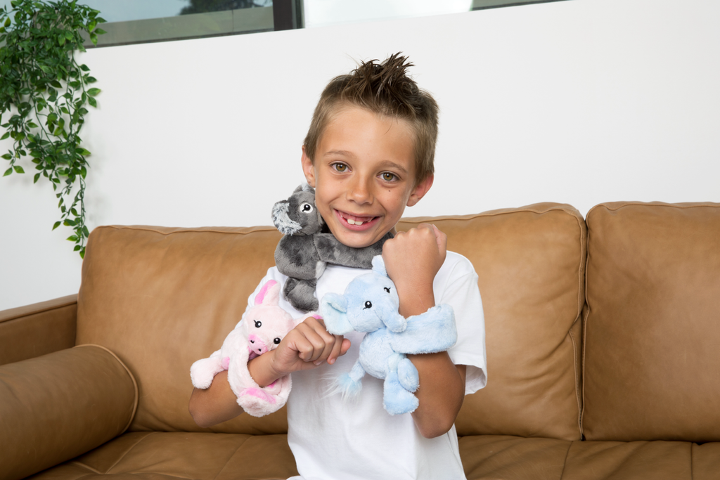 This photo shows a little boy in a white t-shirt smiling as he has three Hug Babies: Pig, Elephant and Koala, wrapped around his wrists and neck.