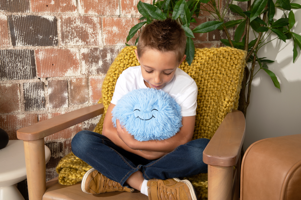 This photo shows a little boy sitting cross-legged in a chair with a mustard yellow blanket, holding the Blue Hug Ball and looking down sweetly at it's "Sweet" expression. 