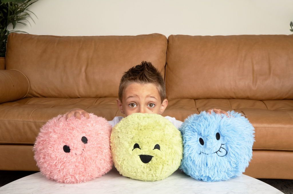 This is a photo of a little boy crouching behind the Pink, Green and Blue Hug Balls so you only see the top of his face and his wide, excited eyes. He's sitting in front of a tan leather couch, and the Hug Balls are on a white round table in front of him. He's holding them in place. 