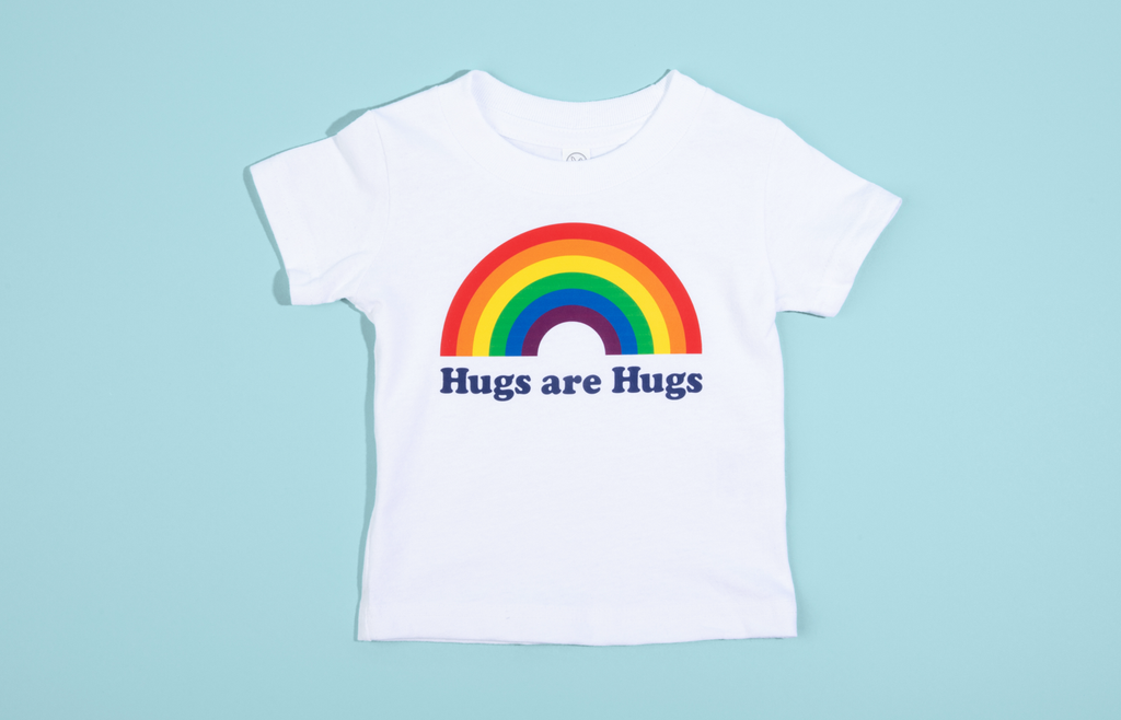 This image shows a white baby sized t-shirt that shows a colorful rainbow with the words Hugs Are Hugs below it in navy lettering . The background is bright teal