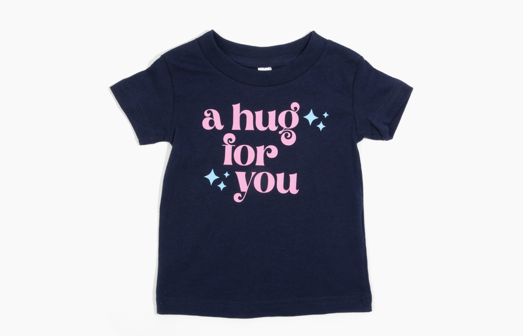 This image shows a navy colored baby size -shirt with the words "A Hug For You" written in pink lettering with a cuople blue stars on the tee also. This is on a white background, 