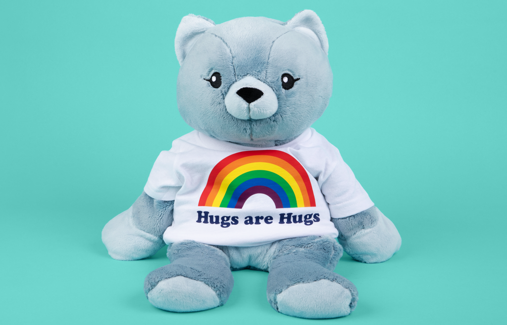 This is an image of Frankie the Cat Hugimal, a light silvery blue colored cat weighted stuffed animal, wearing a white t-shirt that shows a rainbow with the words Hugs Are Hugs below it in navy lettering . The background is bright teal