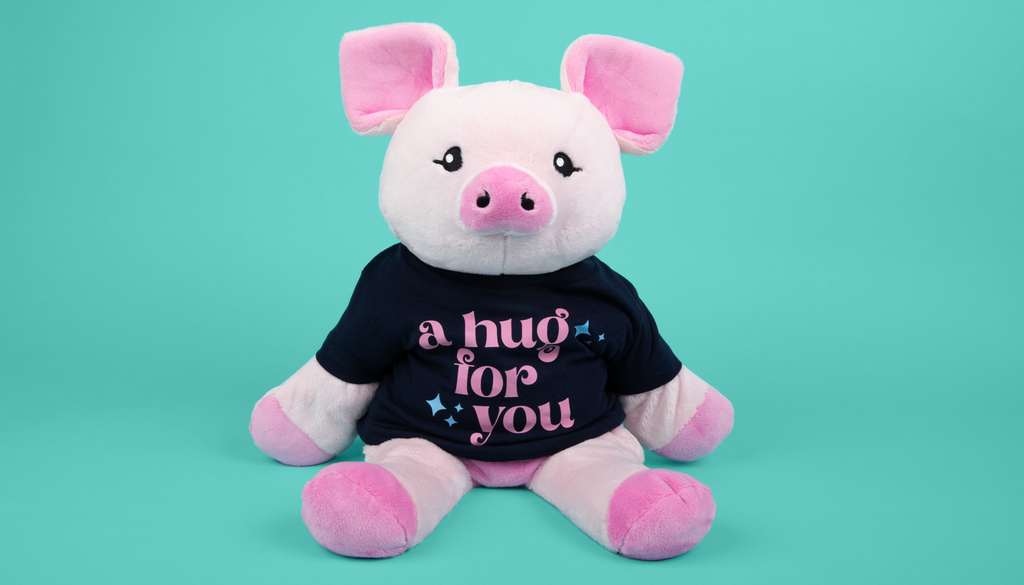 This is an image of Harper the Pig Hugimal, a pink colored piggie weighted stuffed animal, wearing a navy blue t-shirt that says A Hug For You in pink lettering. The background is bright teal.