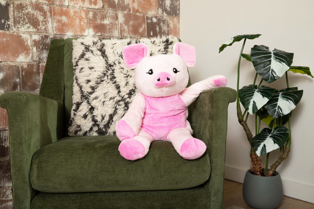 This photo shows a pink pig stuffed animal, Harper the Pig Hugimal, sitting on a green armchair in front of an exposed brick wall with a plant on the floor next to the chair. The pig stuffed animal's arm is relaxing on the arm of the chair how a person's would.