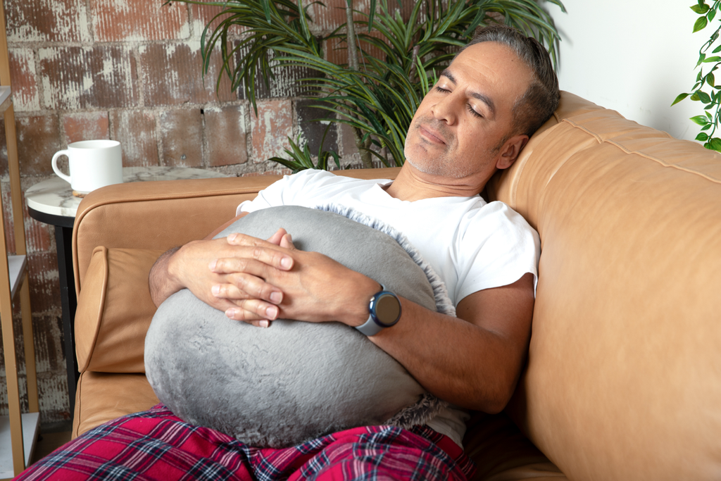 This image shows a dad sleeping while sitting back on a couch, holding a round grey Hug Pillow on his lap