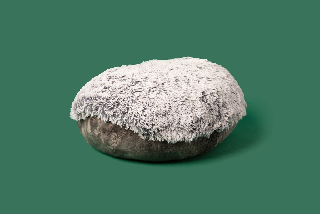 This photo shows the gray round Hug Pillow lying flat on a green background. You can see both the light gray long plush side and the dark gray short plush side, and see the opening to the hand pocket.