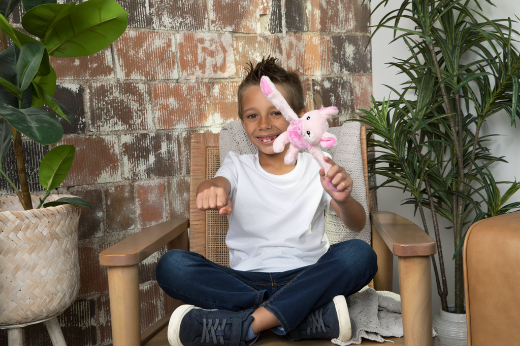 This photo shows a little boy on a chair, wearing a white t-shirt and jeans and black sneakers, about to snap a Pig Hug Baby to his arm. He's smiling and surrounded by plants.