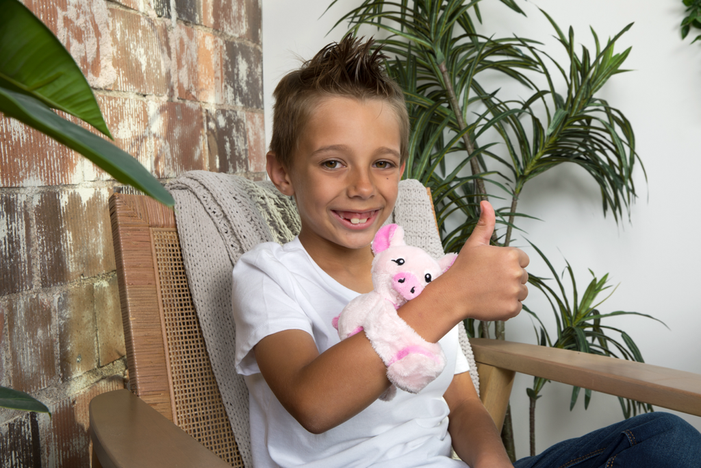 This photo shows a little boy sitting in a chair giving the thumbs up sign and smiling with a Pig Hug Baby on his wrist