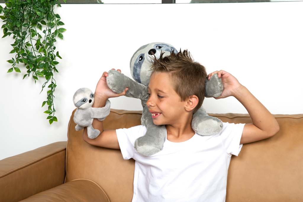 This photo shows a little boy in a white tee shirt smiling, holding a Sam the Sloth Hugimal on his shoulders and looking at a Sloth Hug Baby that's wrapped around his wrist.
