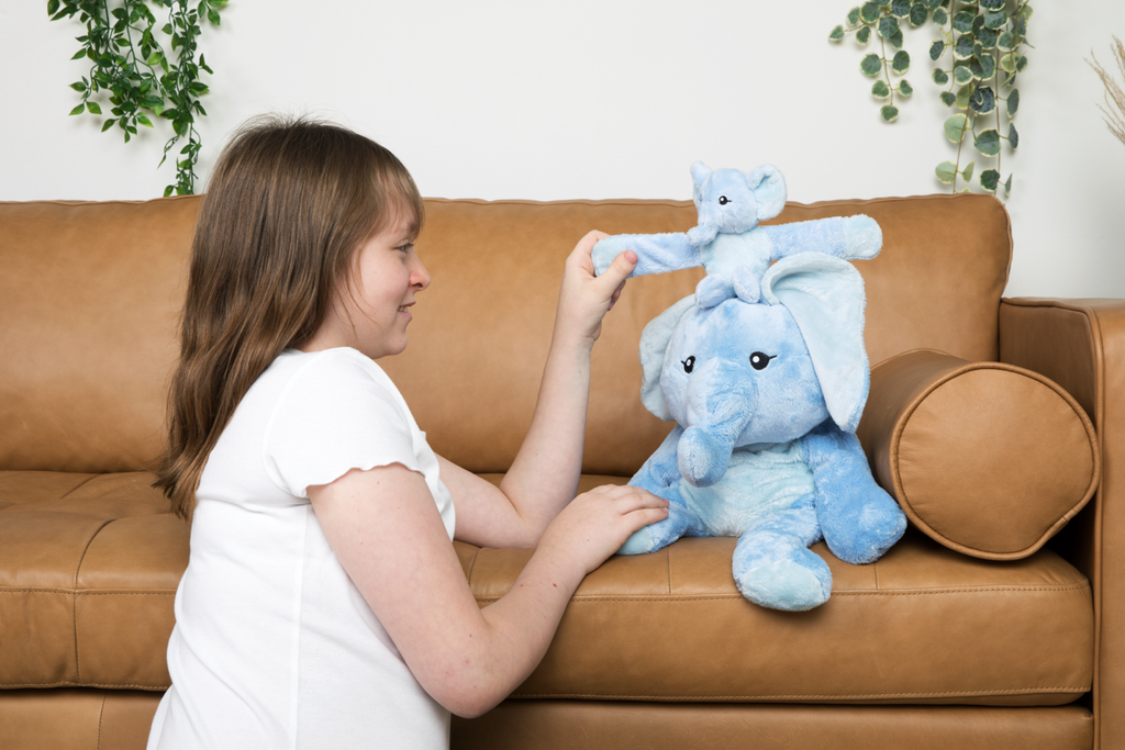 This photo shows a little girl playing with her Elephant Hugimal and Elephant Hug Baby. They are on a tan leather couch, and she is on the floor, smiling.