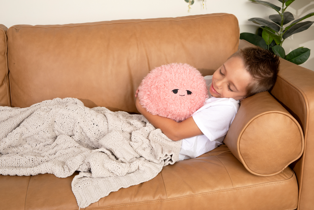This photo shows a little boy reclined on a couch smiling at his Pink Hug Ball, which he's holding. The Pink Hug Ball is showing it's "Sleepy" expression
