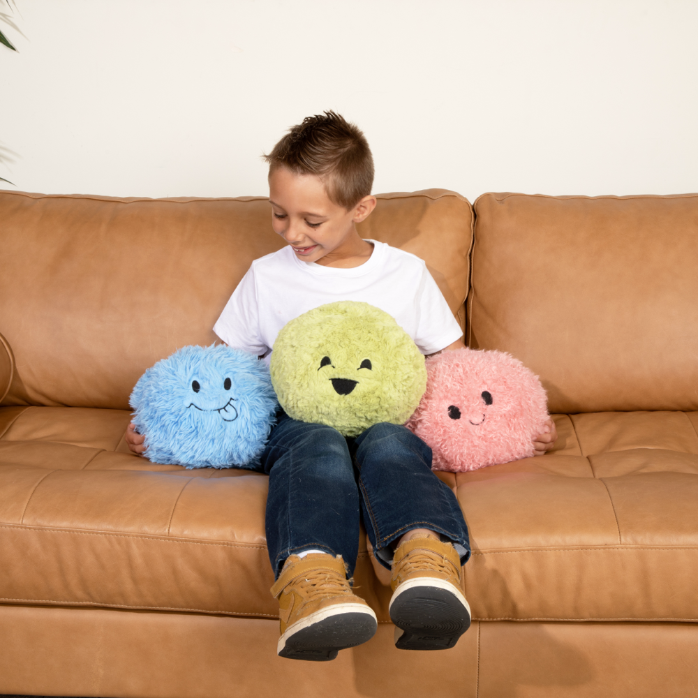 This image shows a little boy (around 8) sitting on a tan couch with a Green Hug Ball on his Lap, a Blue Hug Ball to his right, and a Pink Hug Ball to his right. You can see the smiling faces of each Hug Ball and he is smiling too. He has light brown hair and is wearing a white t-shirt, blue jeans and brown shoes. 