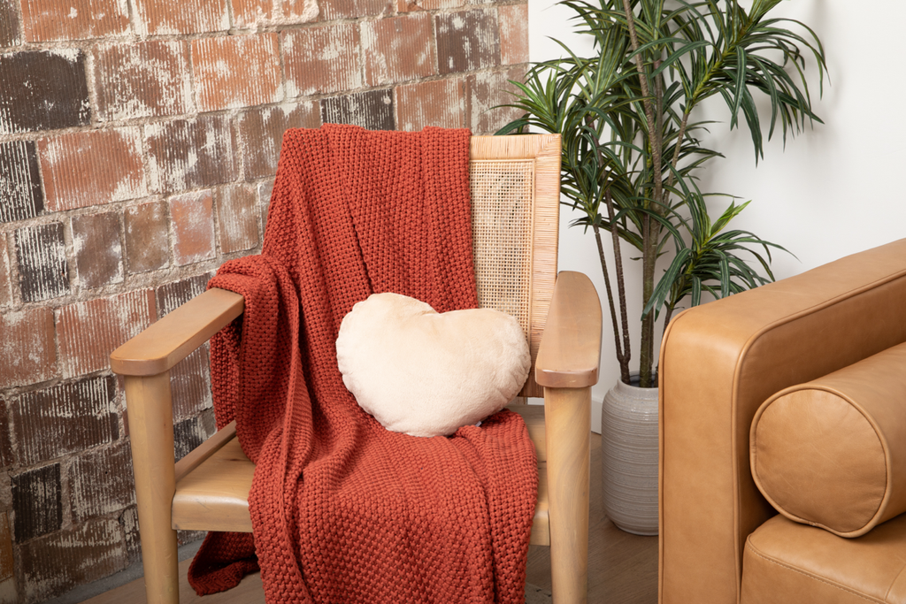 Image shows a Latte Heart to Hug Pillow nestled in a chair with a rust colored blanket