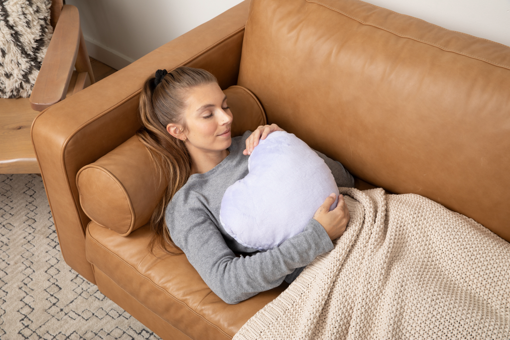 Photo showing young woman lying on couch holding the Lavender Heart to Hug Pillow to her chest, eyes closed with a blanket on her as if taking a nap.
