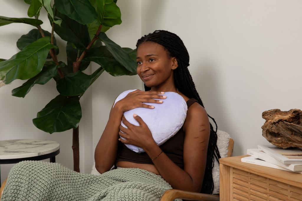 Image shows young woman in chair looking calm and happy, hugging a Lavender Heart to Hug Pillow to her chest, with a plant behind her.