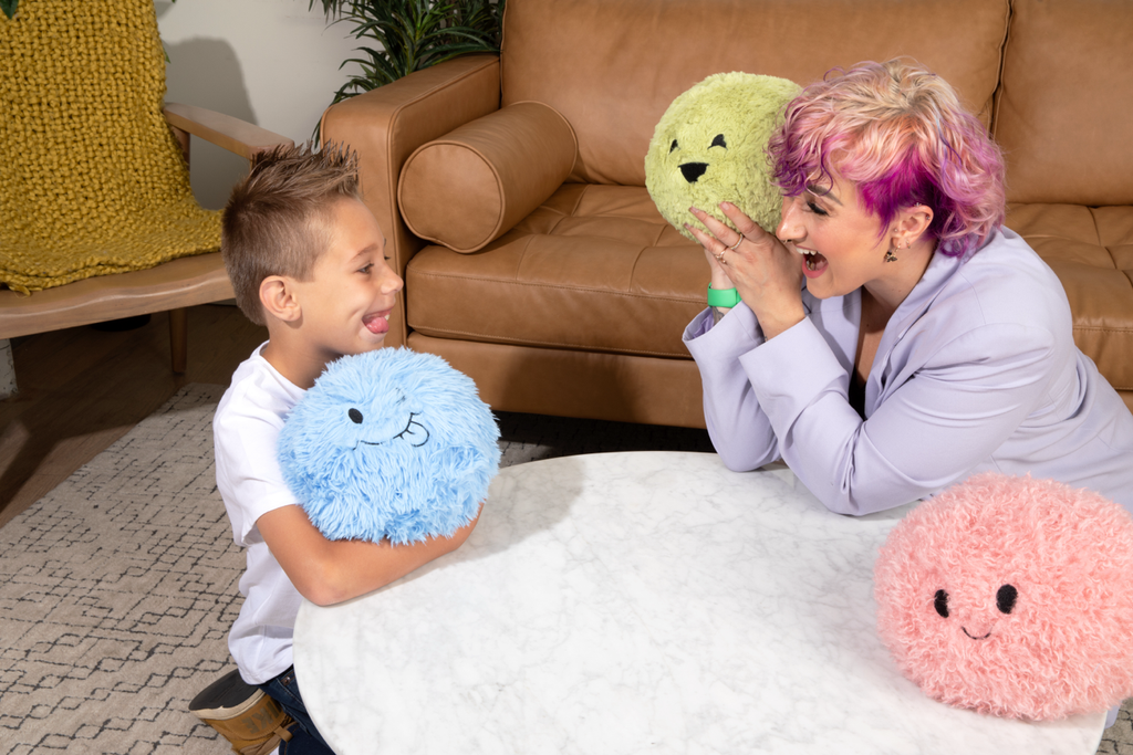An occupational therapist wearing a lavender blazer and a little boy wearomg a white t-shirt smile at each other, sitting on the floor around a white while table. Each is holding a Hug Ball. The little boy is making the same face as his "Silly" Blue Hug Ball. The OT is holding the Green one, and the Pink sits on the table.