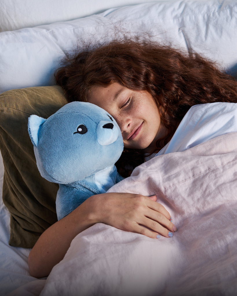This photo shows a little girl with curly red hair tucked in in bed with her eyes closed, hugging Frankie the Cat, a light blue Hugimals weighted stuffed animal.