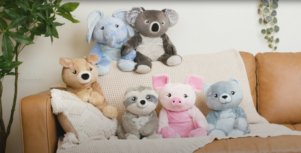 This photo shows six pastel-colored Hugimals weighted plushies sitting together on a tan leather couch draped with a cream blanket. The Hugimals are (clockwise from right bottom): Frankie the Cat, Harper the Pig, Sam the Sloth, Charlie the Puppy, Emory the Elephant, and Quinn the Koala.
