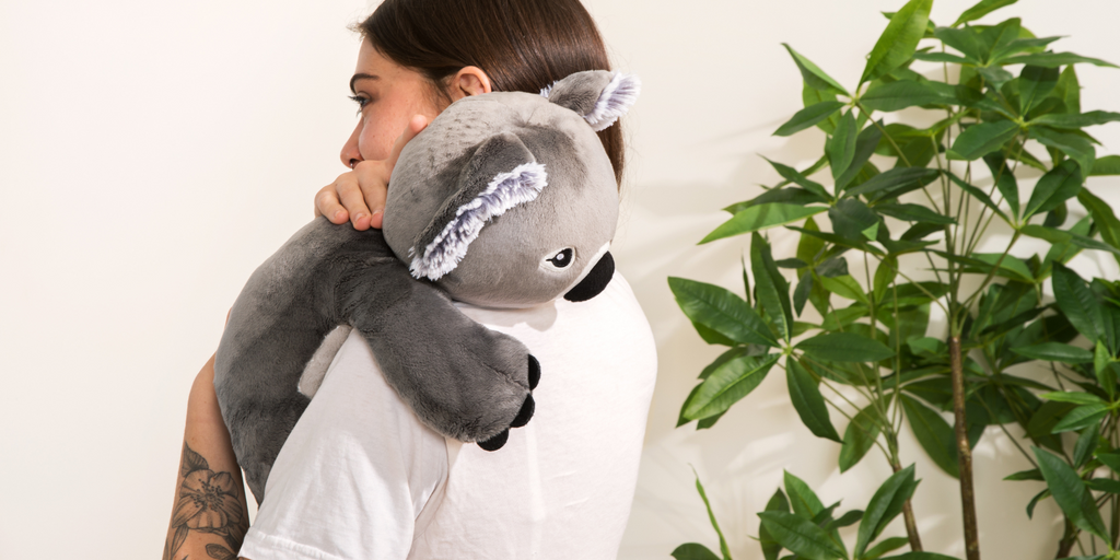 This photo shows a young woman (late teens/early 20s) with dark straight hair, wearing a white t-shirt, and with a floral tattoo showing on her forearm, standing and being hugged by a Quinn the Koala Hugimal. She is standing in profile against a white wall with a tall green plant next to her. We can see the top part of her face, and she looks calm. She is hugging Quinn the Koala back. Quinn is in the position of a baby on her shoulder. 