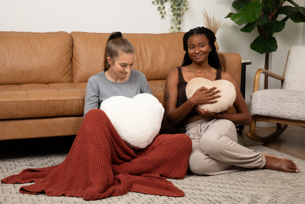 Image showing two young women sitting on the floor holding heart-shaped pillows. One holds the Super Sized Ivory Heart to Hug Pillow, the other holds the Latte Heart to Hug Pillow