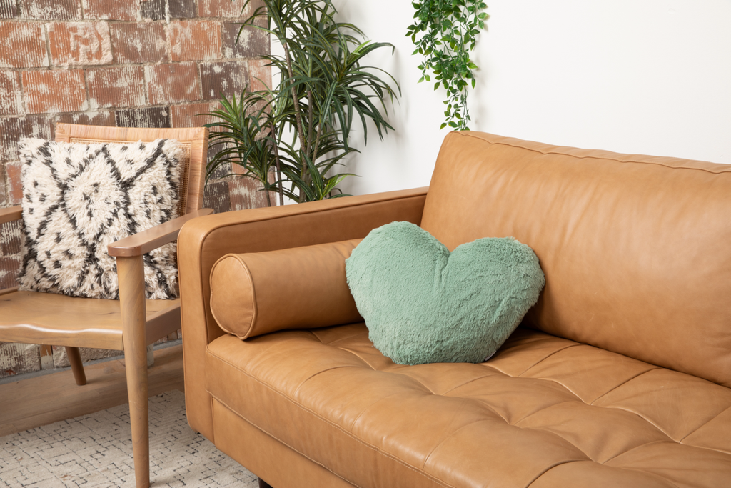 Sage Super Sized Heart to Hug Pillow in the corner of a tan couch, next to some plants and a chair